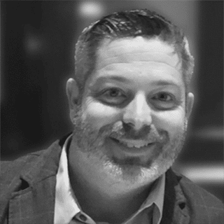 Black and white photo of Ryan Murphy with short dark and gray-colored hair, short trimmed beard, wearing a dark suit jacket and light open-collared dress shirt